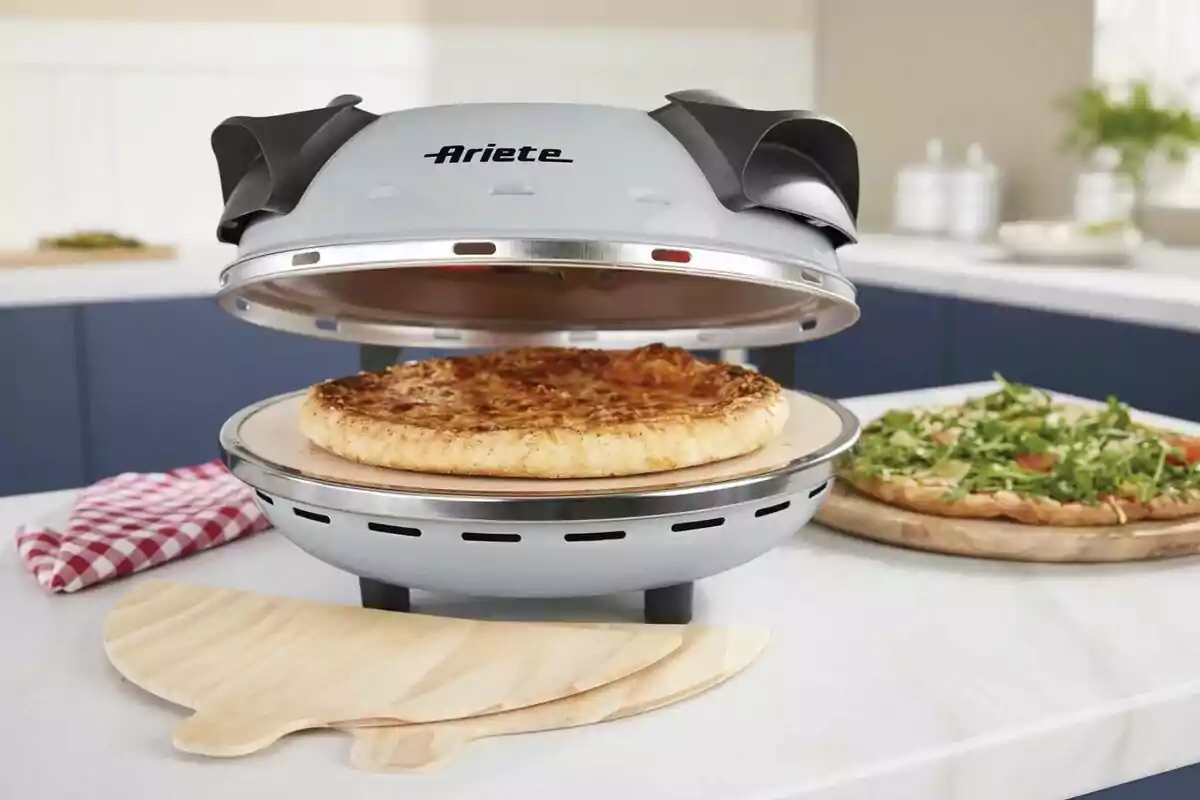 Una pizza cocinándose en un horno eléctrico sobre una encimera de cocina, con una pizza adicional cubierta de rúcula al fondo y un paño de cocina a cuadros rojos y blancos al lado. Una pizza cocinándose en un horno eléctrico sobre una encimera de cocina, con una pizza adicional cubierta de rúcula al fondo y un paño de cocina a cuadros rojos y blancos al lado.