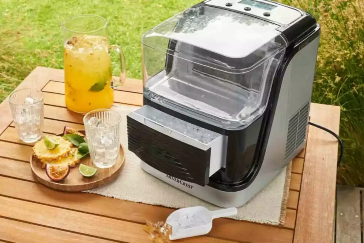 Máquina de hacer hielo sobre una mesa de madera junto a una jarra de bebida amarilla con hielo, dos vasos, una charola con rodajas de piña, lima y maracuyá, y un cucharón de plástico con hielo. Máquina de hacer hielo sobre una mesa de madera junto a una jarra de bebida amarilla con hielo, dos vasos, una charola con rodajas de piña, lima y maracuyá, y un cucharón de plástico con hielo.
