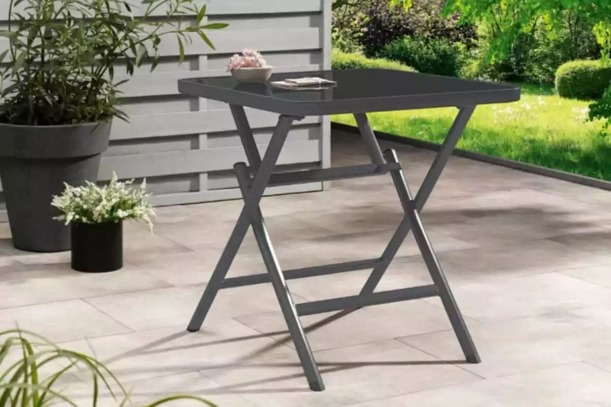 Una mesa plegable de color oscuro está situada en una terraza con piso de baldosas, rodeada de plantas en macetas y un jardín verde al fondo. Una mesa plegable de color oscuro está situada en una terraza con piso de baldosas, rodeada de plantas en macetas y un jardín verde al fondo.