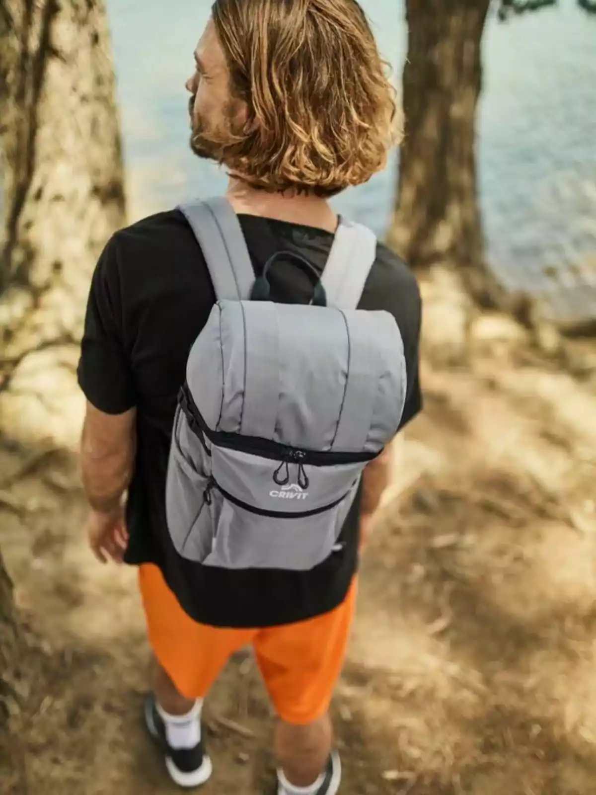 Un hombre con cabello largo y barba lleva una mochila gris mientras camina por un sendero boscoso cerca de un lago, vistiendo una camiseta negra y pantalones cortos naranjas. Un hombre con cabello largo y barba lleva una mochila gris mientras camina por un sendero boscoso cerca de un lago, vistiendo una camiseta negra y pantalones cortos naranjas.