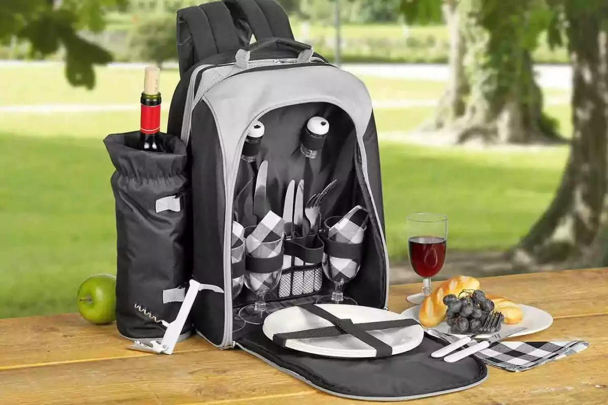 Una mochila de picnic abierta sobre una mesa de madera en un parque, mostrando su contenido que incluye cubiertos, copas, platos y servilletas, con una botella de vino en un compartimento lateral, un sacacorchos, una manzana verde y un plato con uvas, panecillos y una copa de vino tinto. Una mochila de picnic abierta sobre una mesa de madera en un parque, mostrando su contenido que incluye cubiertos, copas, platos y servilletas, con una botella de vino en un compartimento lateral, un sacacorchos, una manzana verde y un plato con uvas, panecillos y una copa de vino tinto.