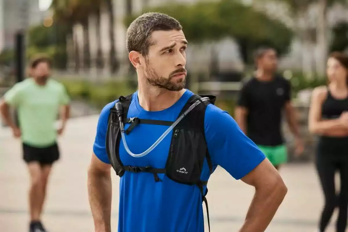 Hombre con camiseta azul y mochila de hidratación en un entorno al aire libre con personas en el fondo. Hombre con camiseta azul y mochila de hidratación en un entorno al aire libre con personas en el fondo.