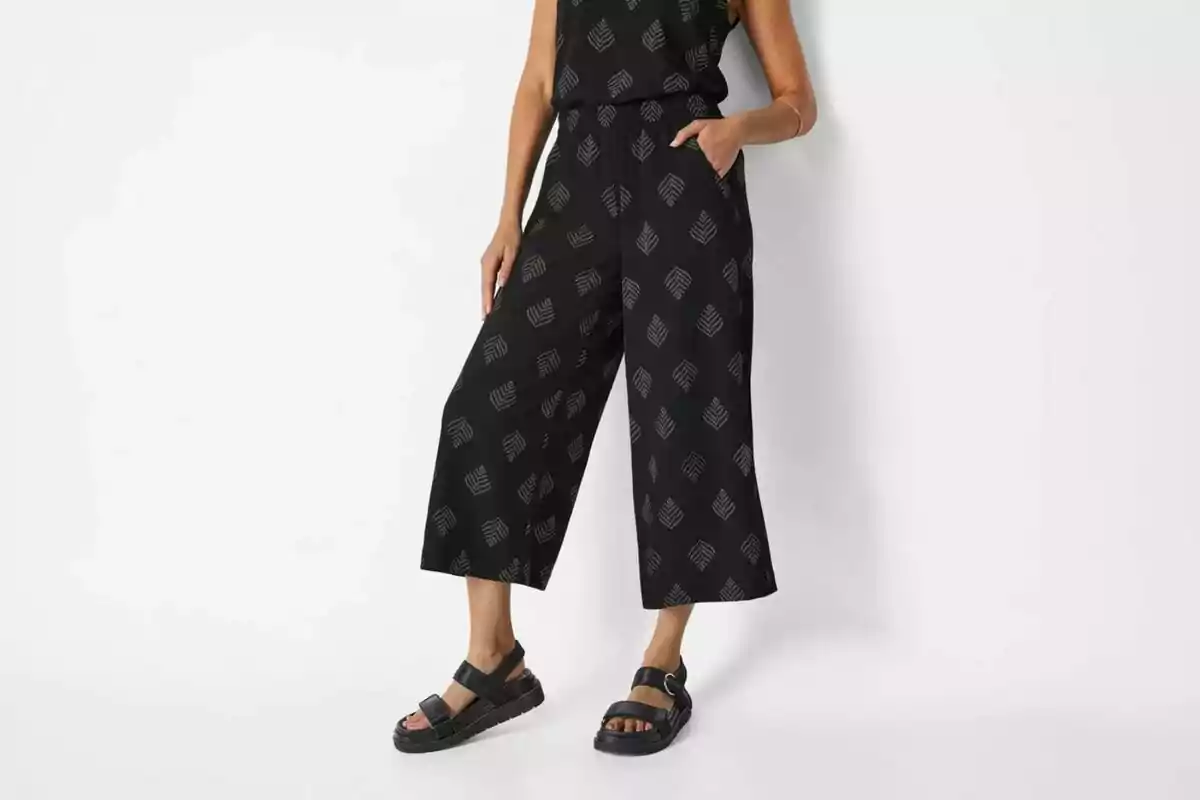 Mujer usando un jumpsuit negro con estampado de hojas y sandalias negras sobre fondo blanco Mujer usando un jumpsuit negro con estampado de hojas y sandalias negras sobre fondo blanco