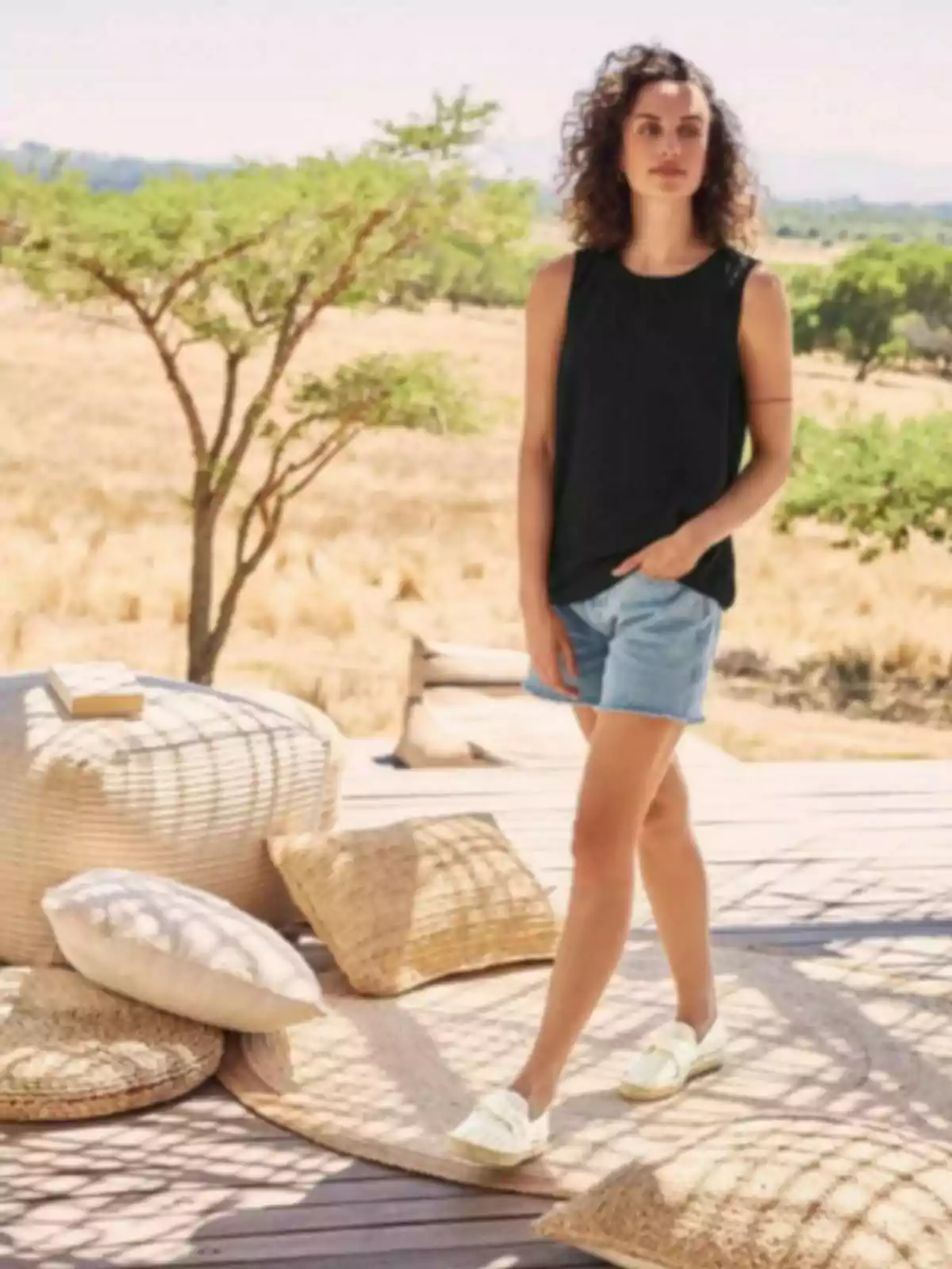 Mujer de cabello rizado con blusa negra y shorts de mezclilla posando al aire libre sobre una terraza de madera decorada con cojines y alfombras tejidas en un entorno natural y soleado Mujer de cabello rizado con blusa negra y shorts de mezclilla posando al aire libre sobre una terraza de madera decorada con cojines y alfombras tejidas en un entorno natural y soleado