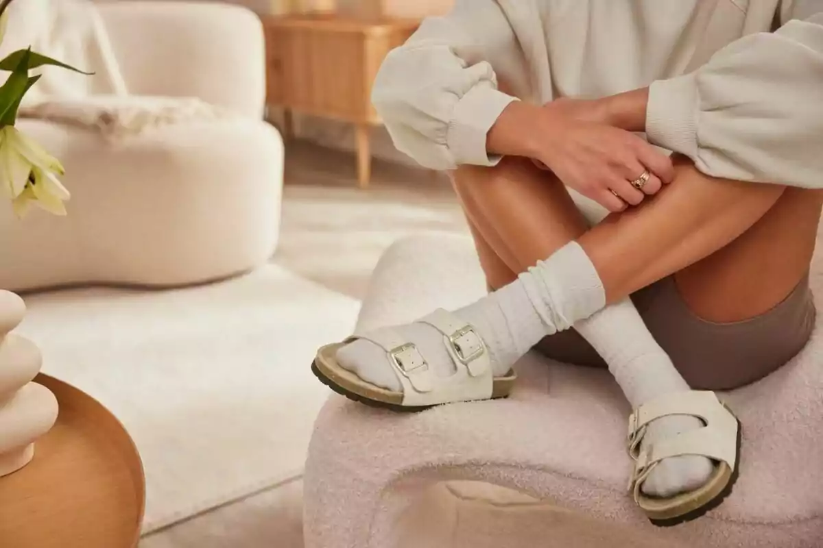 Persona sentada en una silla moderna con sandalias blancas y calcetas, en una sala de estar de tonos claros y decoración minimalista Persona sentada en una silla moderna con sandalias blancas y calcetas, en una sala de estar de tonos claros y decoración minimalista