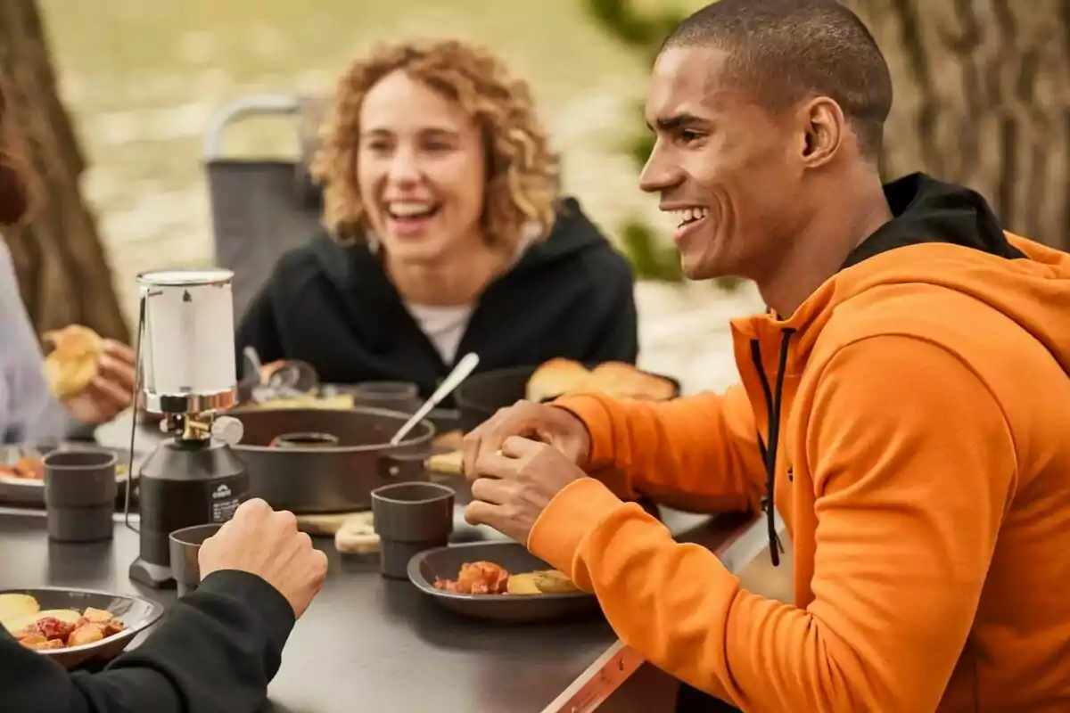 Un grupo de personas sonríe mientras disfruta de una comida al aire libre alrededor de una mesa con platos y tazas. Un grupo de personas sonríe mientras disfruta de una comida al aire libre alrededor de una mesa con platos y tazas.