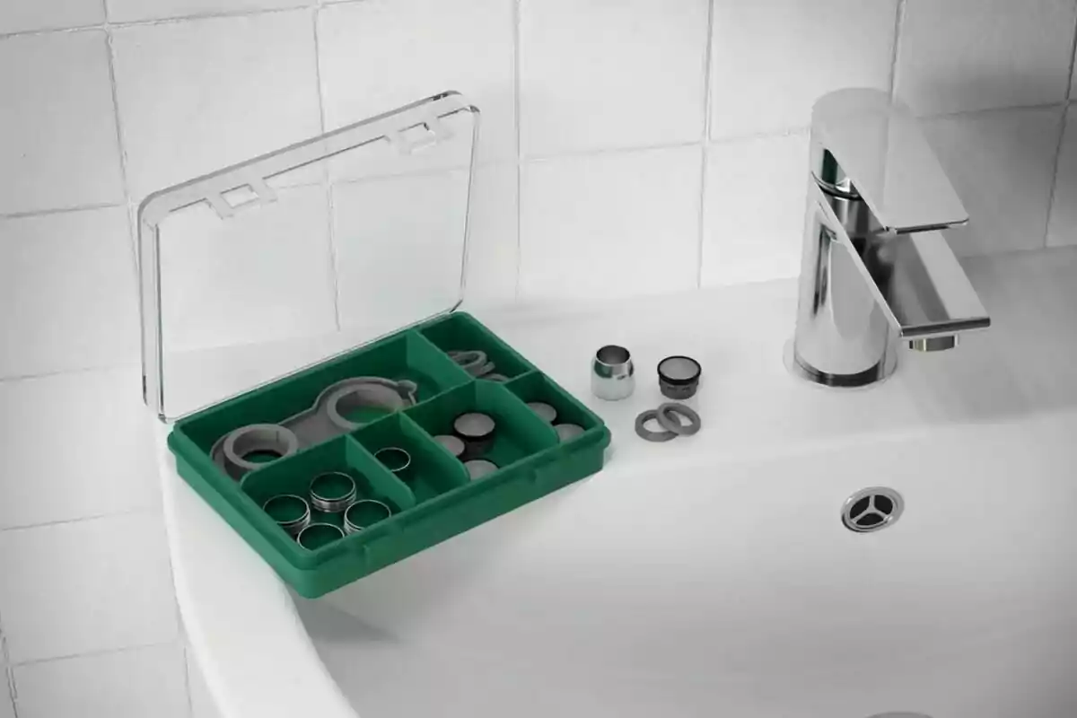 Caja verde con compartimentos que contiene piezas y herramientas pequeñas sobre un lavabo blanco junto a una llave de baño plateada Caja verde con compartimentos que contiene piezas y herramientas pequeñas sobre un lavabo blanco junto a una llave de baño plateada