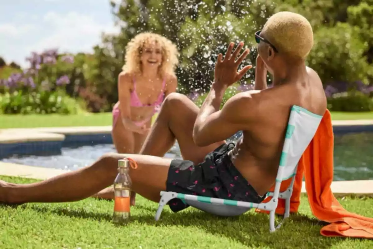 Dos personas se divierten junto a una piscina en un día soleado una de ellas está sentada en una silla baja con una toalla naranja y una botella de agua mientras la otra persona le salpica agua y sonríe Dos personas se divierten junto a una piscina en un día soleado una de ellas está sentada en una silla baja con una toalla naranja y una botella de agua mientras la otra persona le salpica agua y sonríe