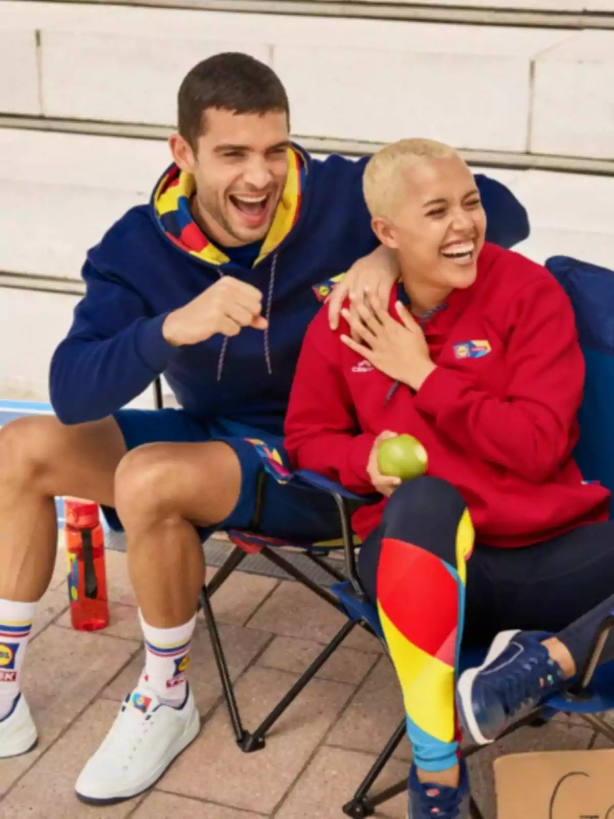 Dos personas sentadas en sillas plegables, riendo y disfrutando de un momento al aire libre, una de ellas sostiene una manzana verde. Dos personas sentadas en sillas plegables, riendo y disfrutando de un momento al aire libre, una de ellas sostiene una manzana verde.