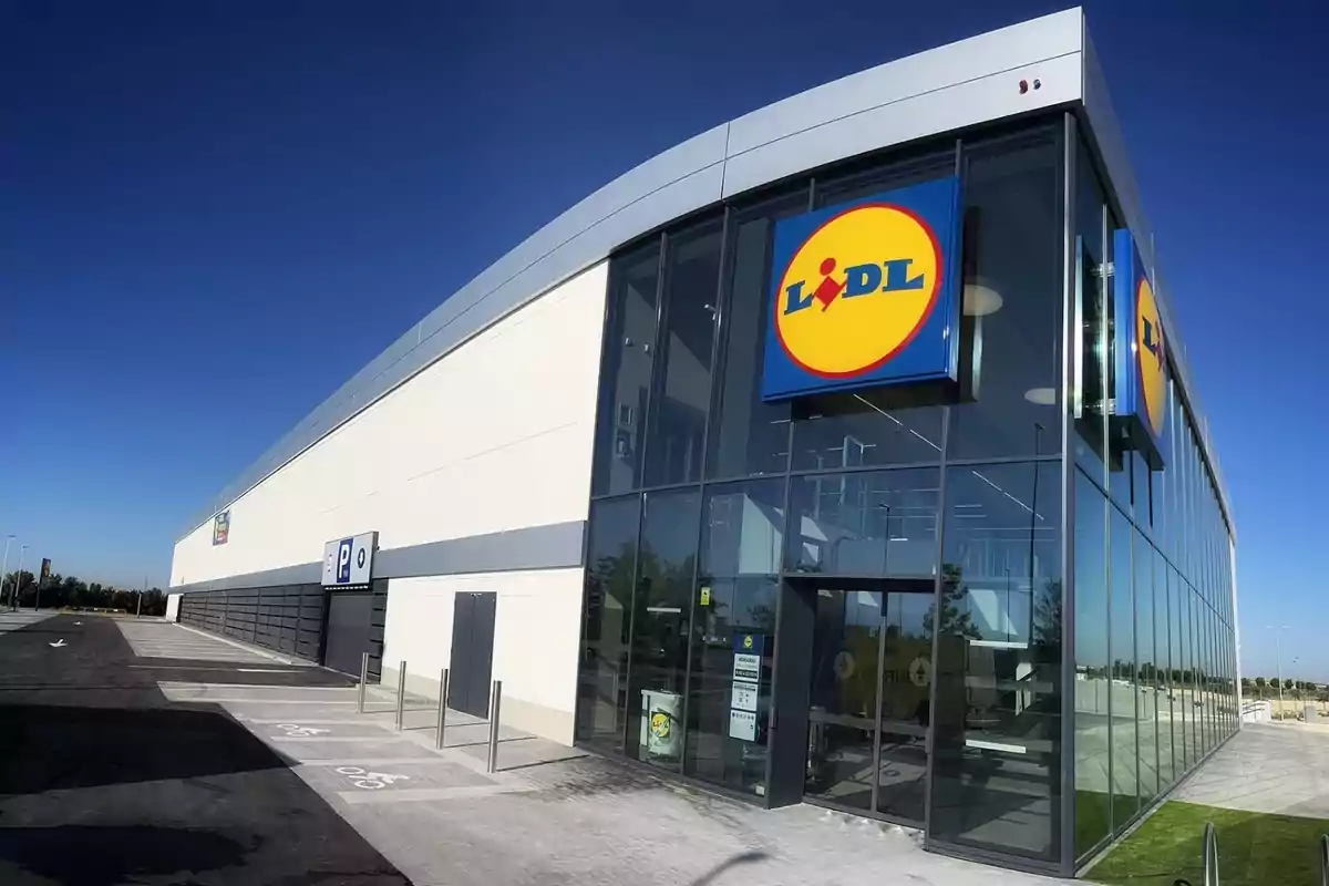 Lidl grande, de extrarradio, con paredes acristaladas por la parte de la tienda y, a la izquierda, la entrada al parking subterráneo con pared de hormigón. Lidl grande, de extrarradio, con paredes acristaladas por la parte de la tienda y, a la izquierda, la entrada al parking subterráneo con pared de hormigón.