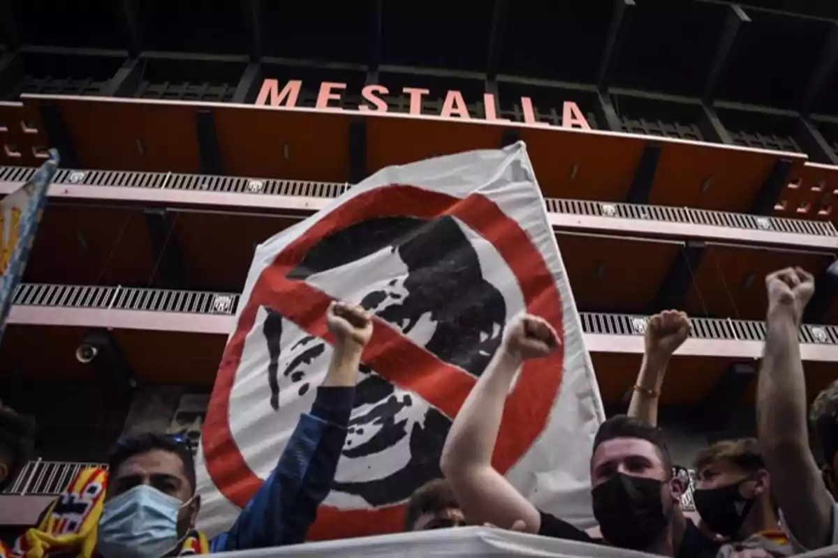 Personas protestando frente al estadio Mestalla con un cartel que tiene un símbolo de prohibición sobre un rostro.