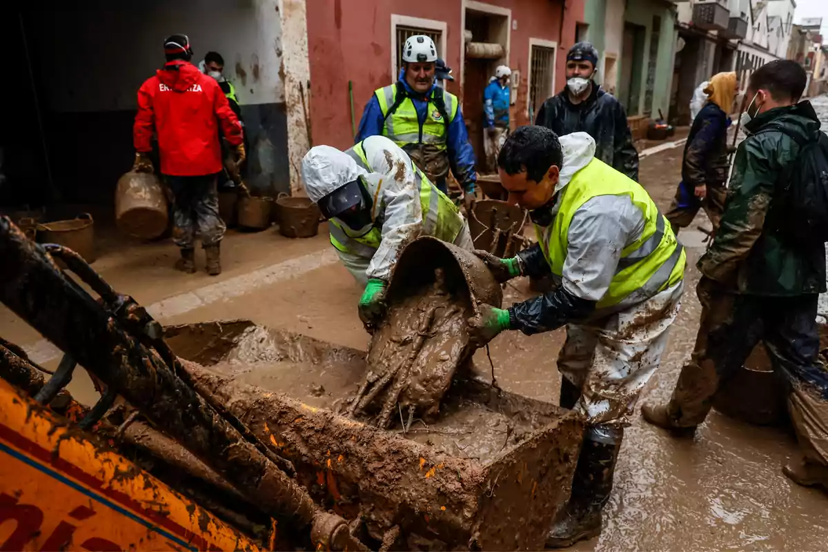 Personas con equipo de protección y chalecos reflectantes limpian lodo en una calle después de una inundación.