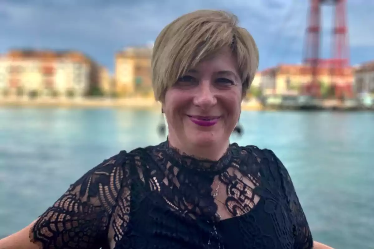 Mujer sonriente con cabello corto rubio y labial rosa, vestida con blusa negra de encaje, posando frente a un río con edificios y un puente al fondo Mujer sonriente con cabello corto rubio y labial rosa, vestida con blusa negra de encaje, posando frente a un río con edificios y un puente al fondo
