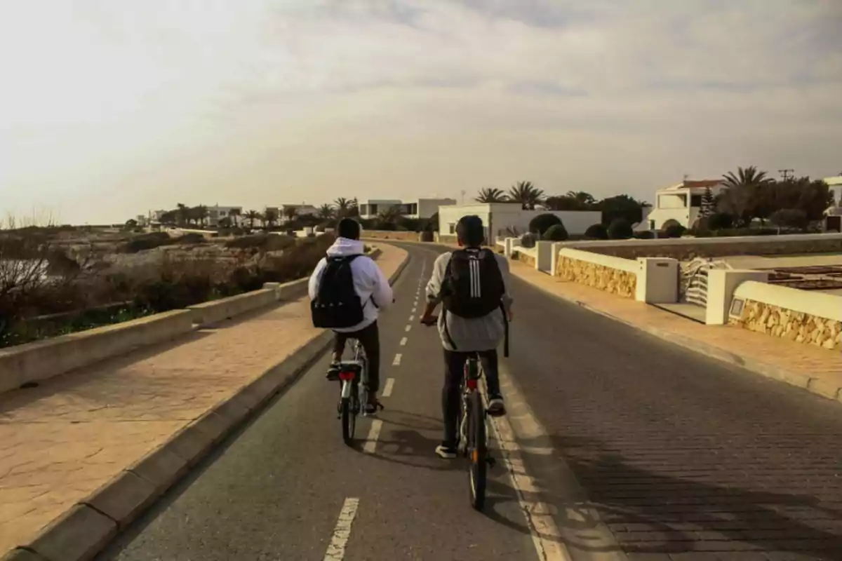 Dos personas con mochilas montan bicicletas por una calle tranquila rodeada de casas y vegetación bajo un cielo nublado.