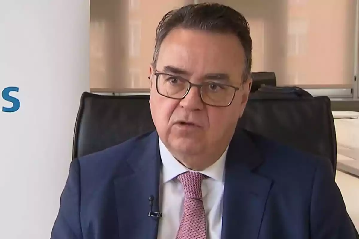 Un hombre con gafas y traje azul está sentado en una oficina, hablando frente a una cámara. Un hombre con gafas y traje azul está sentado en una oficina, hablando frente a una cámara.