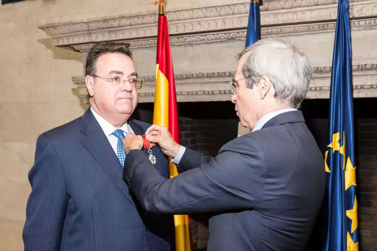 Un hombre recibe una medalla de honor de otro hombre en una ceremonia formal, con banderas de fondo.
