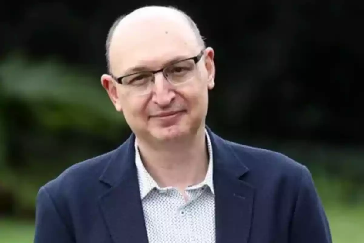 Un hombre con gafas y chaqueta oscura sonríe ligeramente al aire libre. Un hombre con gafas y chaqueta oscura sonríe ligeramente al aire libre.