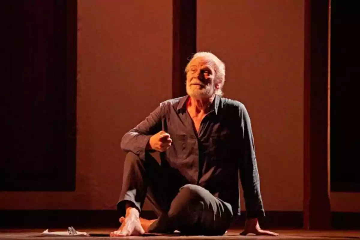 Un hombre mayor con barba y cabello canoso, sentado descalzo en el suelo de un escenario, con una expresión pensativa y una hoja de papel a su lado.