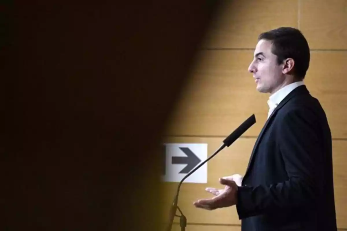 Un hombre de perfil hablando en un micrófono con un fondo de madera y una flecha en la pared. Un hombre de perfil hablando en un micrófono con un fondo de madera y una flecha en la pared.