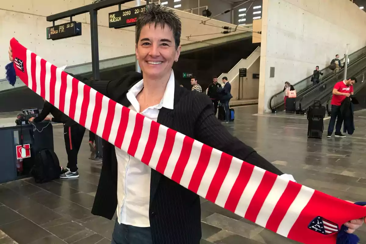 Una persona sonriente sostiene una bufanda de rayas rojas y blancas en una estación de tren. Una persona sonriente sostiene una bufanda de rayas rojas y blancas en una estación de tren.