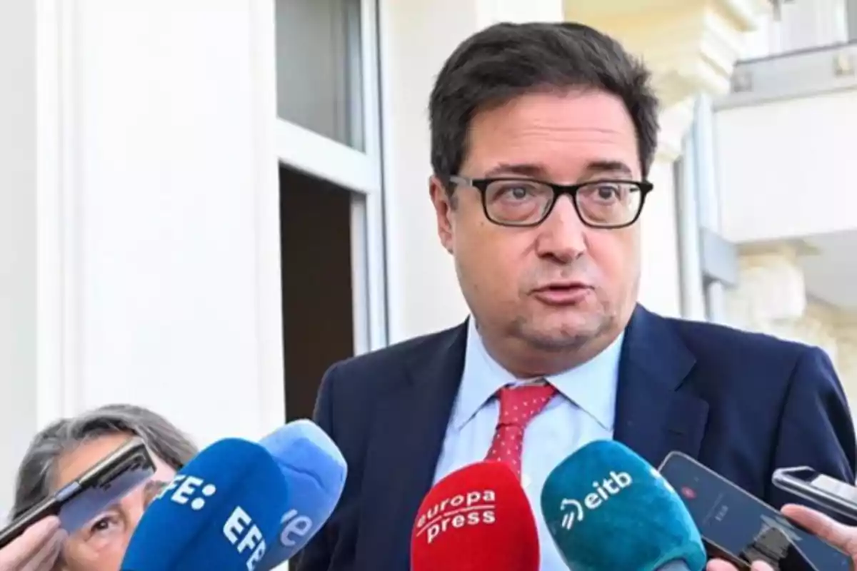 Un hombre con gafas y traje oscuro habla frente a varios micrófonos de diferentes medios de comunicación.