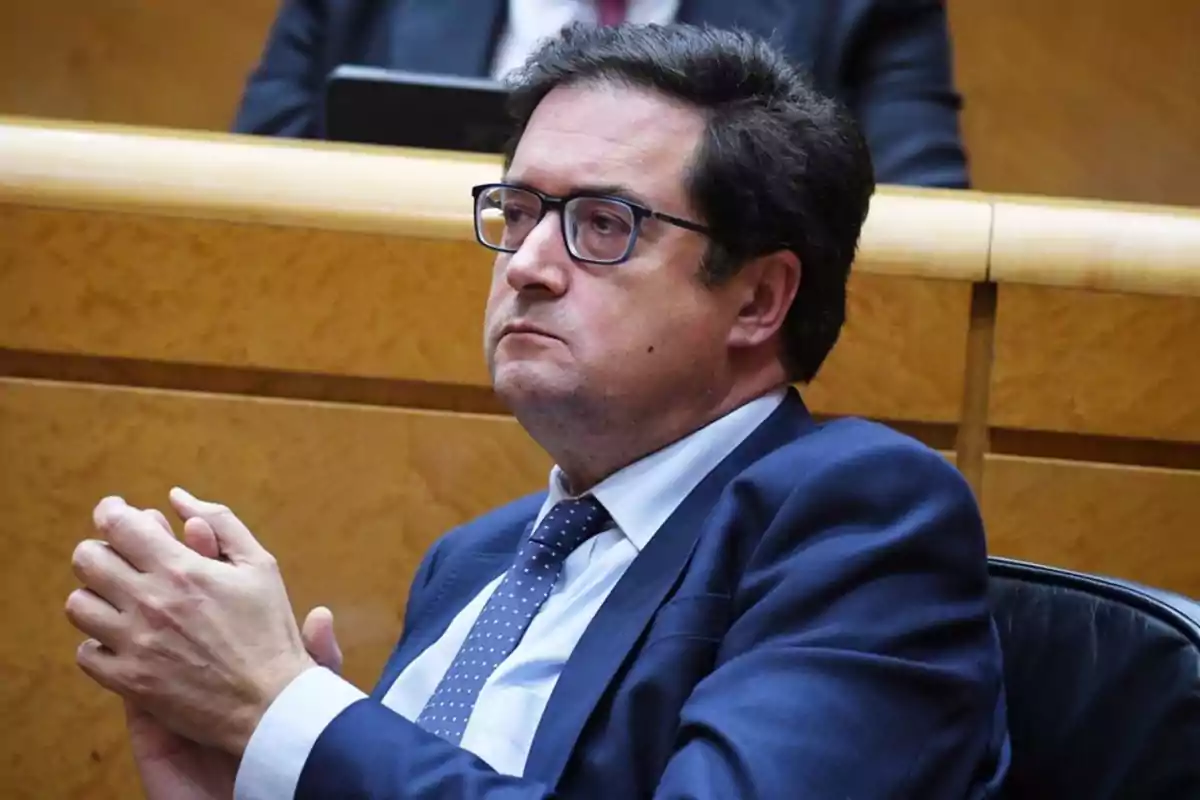 Hombre con gafas y traje azul sentado en un entorno formal.