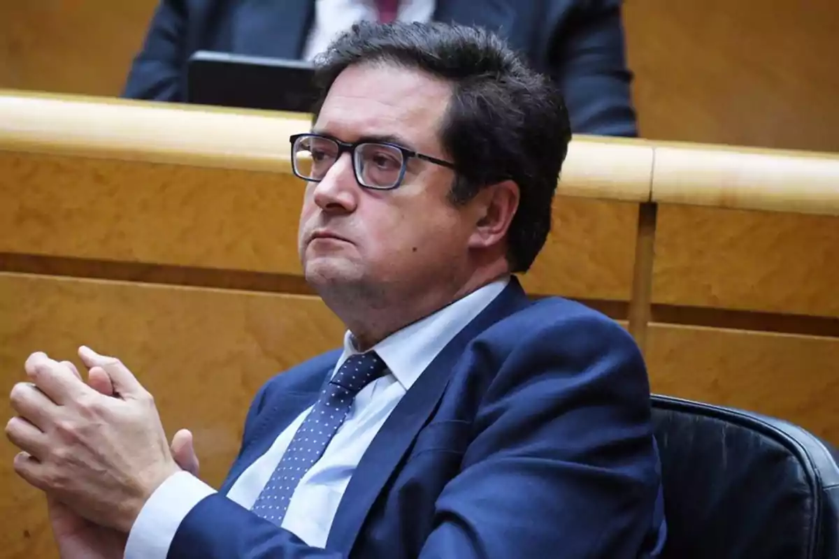 Un hombre con gafas y traje azul está sentado en un entorno formal, mirando hacia adelante con expresión seria. Un hombre con gafas y traje azul está sentado en un entorno formal, mirando hacia adelante con expresión seria.