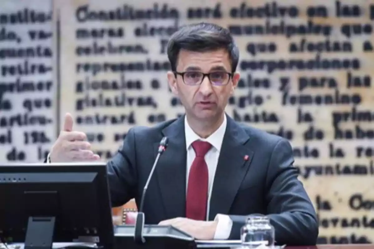 Un hombre con traje y corbata roja habla en un micrófono en una sala con un fondo de texto borroso. Un hombre con traje y corbata roja habla en un micrófono en una sala con un fondo de texto borroso.