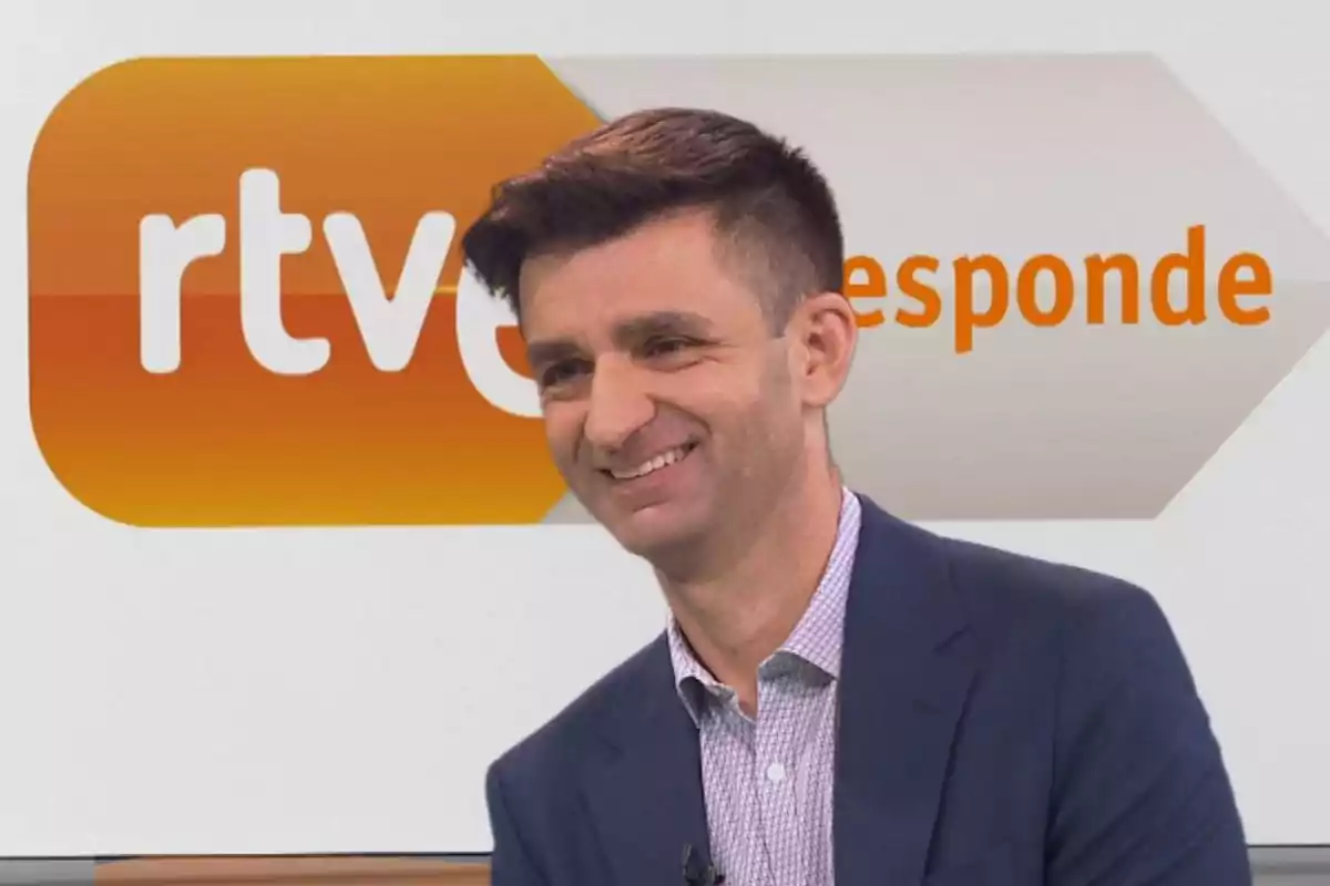 Hombre sonriente con saco azul y camisa clara frente a un fondo con el logo de rtve responde Hombre sonriente con saco azul y camisa clara frente a un fondo con el logo de rtve responde