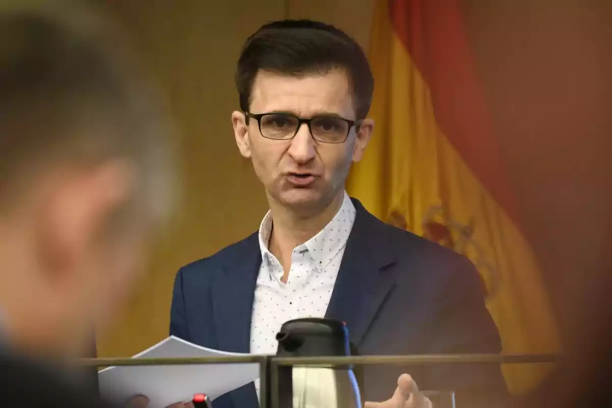 Hombre con gafas y chaqueta azul hablando en público con una bandera de España al fondo Hombre con gafas y chaqueta azul hablando en público con una bandera de España al fondo