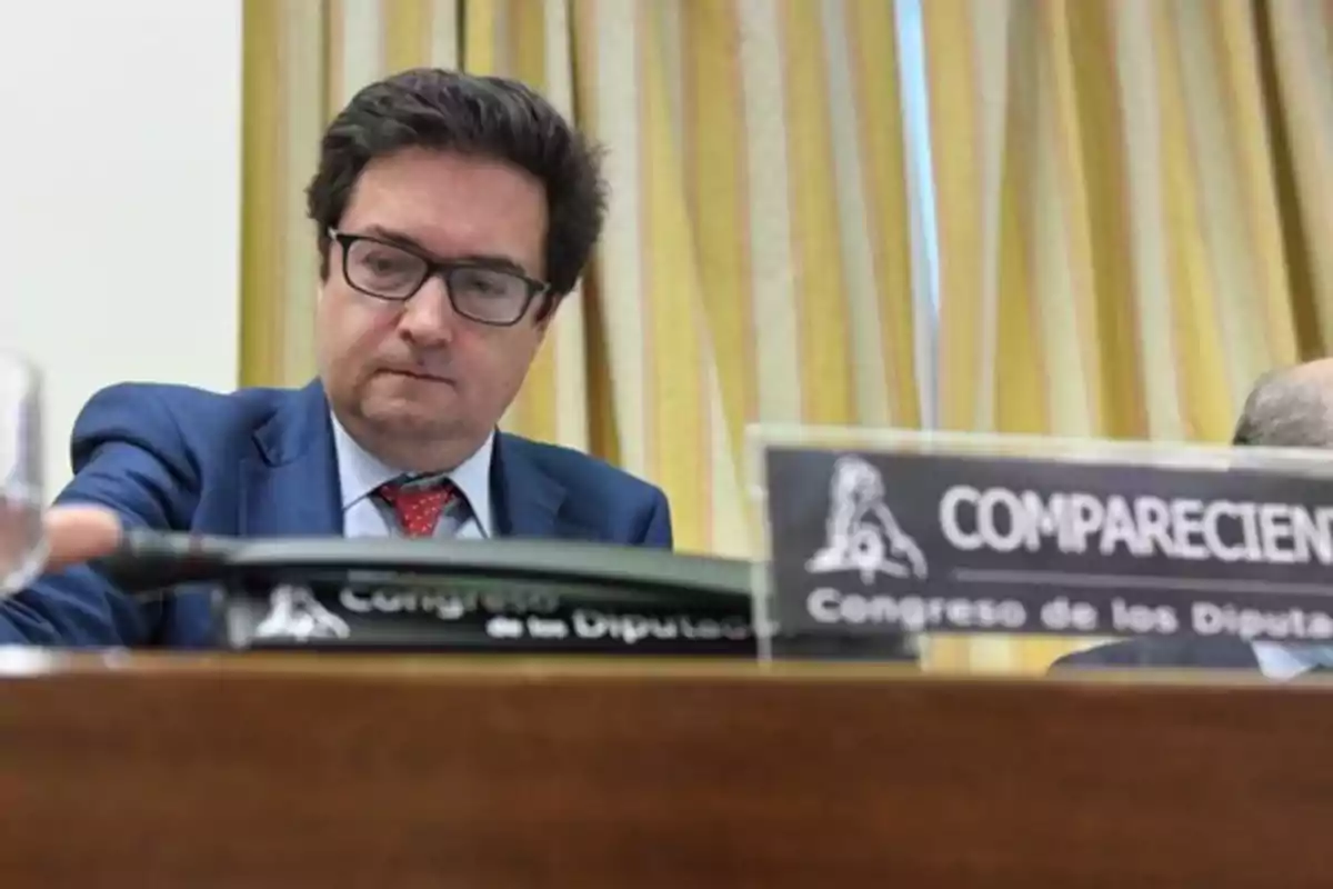 Un hombre con gafas y traje azul sentado en una mesa con un cartel que dice "COMPARECIENTE Congreso de los Diputados".