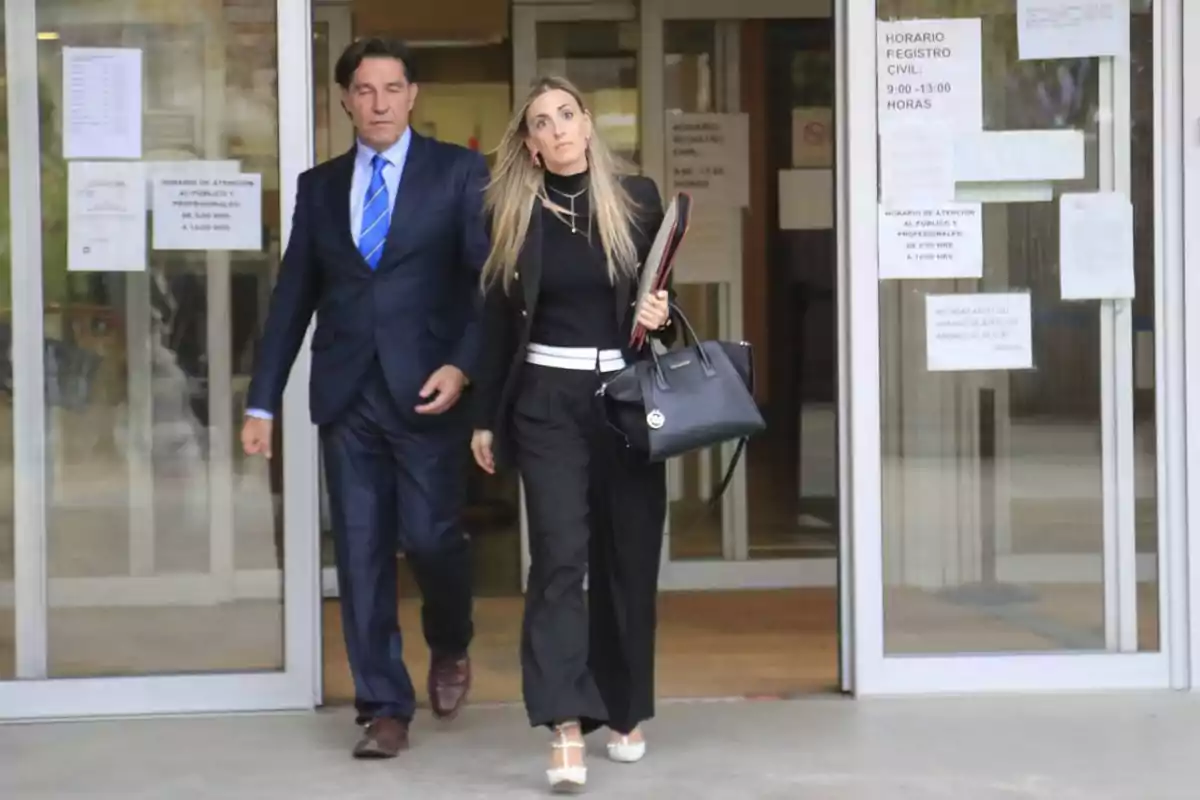 Dos personas bien vestidas salen de un edificio con puertas de vidrio, una mujer lleva una bolsa negra y documentos mientras un hombre con traje y corbata azul camina a su lado.