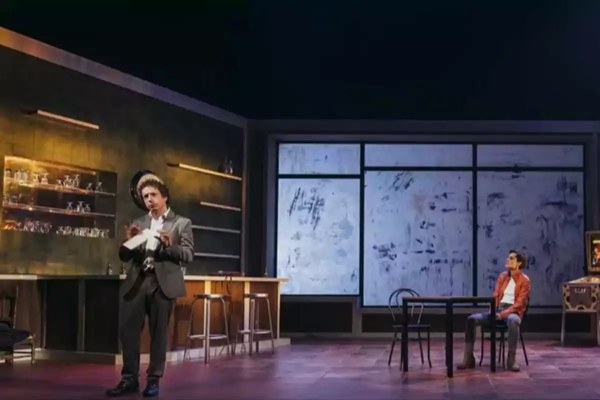 Dos actores en un escenario de teatro, uno de pie junto a una barra y el otro sentado en una mesa.