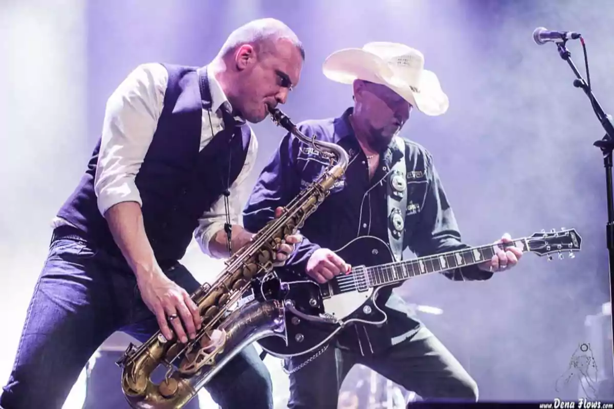 Dos músicos tocando en el escenario, uno con saxofón y otro con guitarra eléctrica, bajo luces de colores.