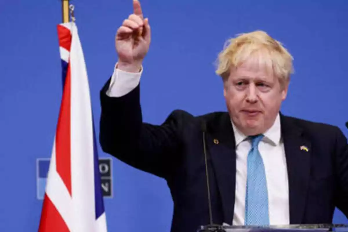 Hombre de traje y corbata azul levantando la mano junto a una bandera del Reino Unido.