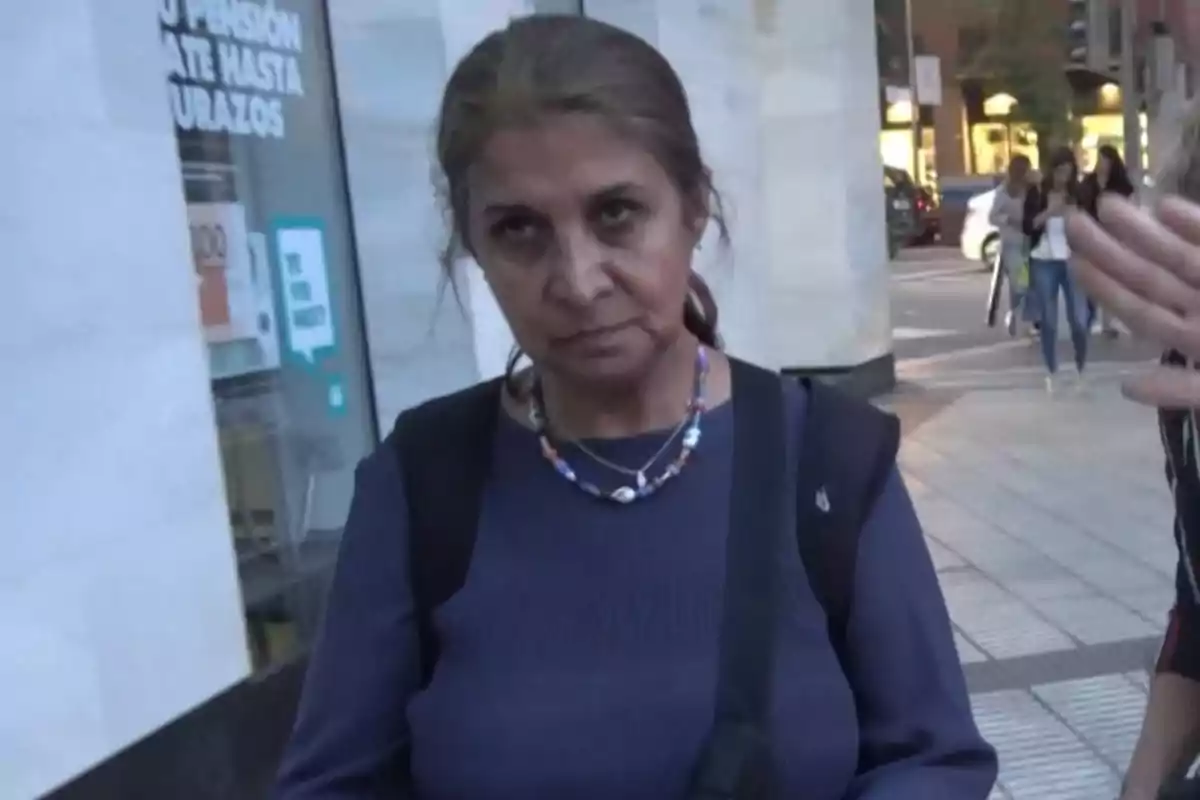 Una mujer de pie en una calle urbana con expresión seria, lleva un collar colorido y una mochila.