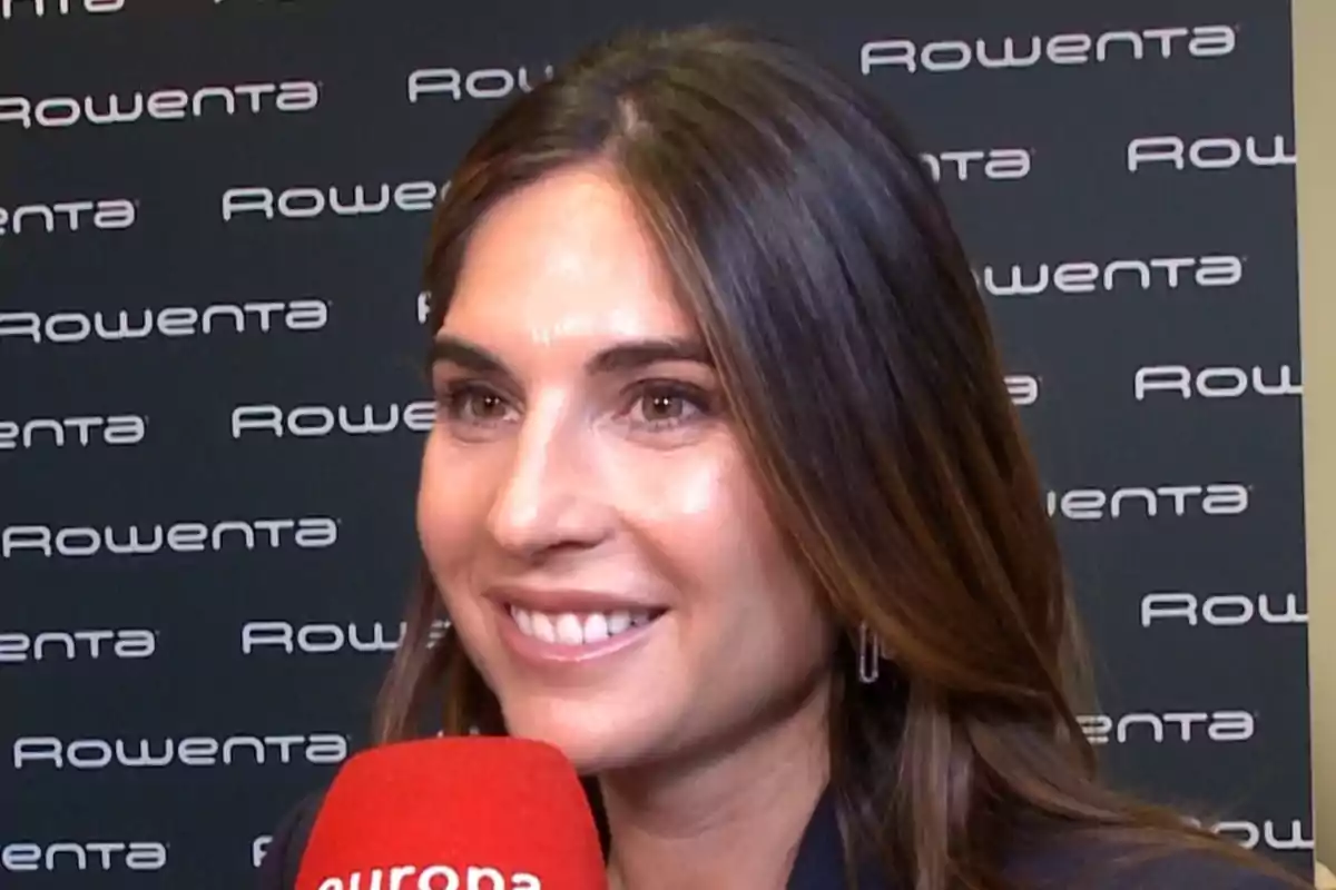 Lourdes Montes sonriendo sostiene un micrófono rojo frente a un fondo con el logotipo de Rowenta repetido. Lourdes Montes sonriendo sostiene un micrófono rojo frente a un fondo con el logotipo de Rowenta repetido.
