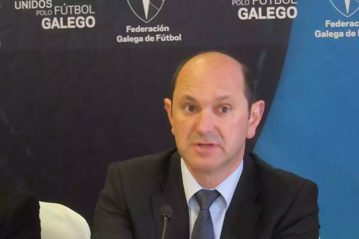 Un hombre en traje hablando frente a un micrófono con un fondo que muestra el logotipo de la Federación Galega de Fútbol.