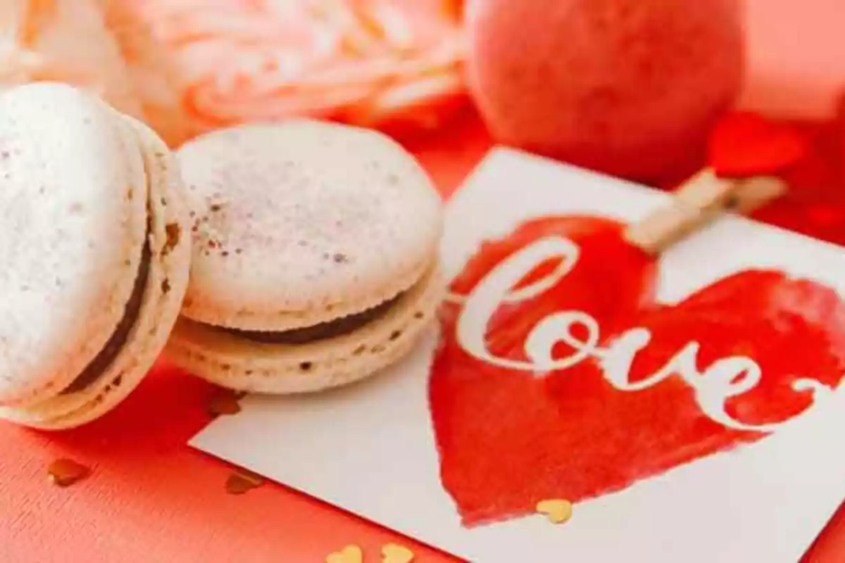 Macarons blancos junto a una tarjeta con un corazón rojo y la palabra "love" sobre una superficie roja.