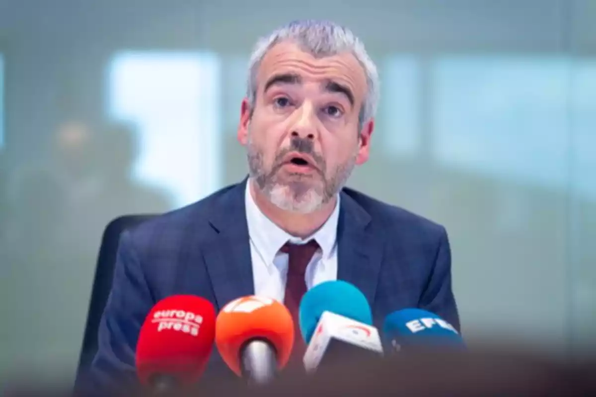 Hombre de cabello canoso y barba hablando en una conferencia de prensa con varios micrófonos de diferentes medios frente a él Hombre de cabello canoso y barba hablando en una conferencia de prensa con varios micrófonos de diferentes medios frente a él