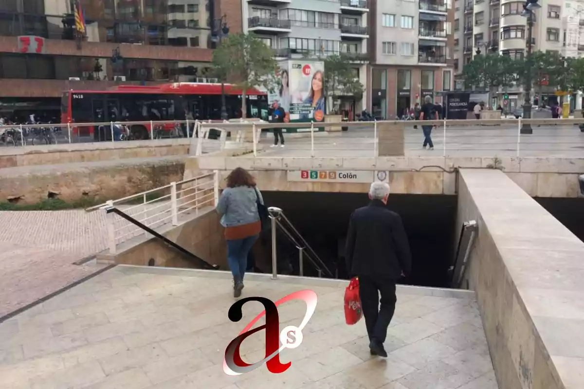 Personas descendiendo hacia la entrada de una estación de metro en una ciudad, con un autobús rojo y edificios al fondo.