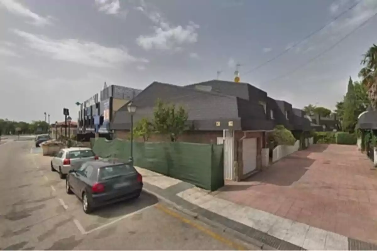 Calle con coches estacionados junto a una fila de casas con techos oscuros y una valla verde. Calle con coches estacionados junto a una fila de casas con techos oscuros y una valla verde.