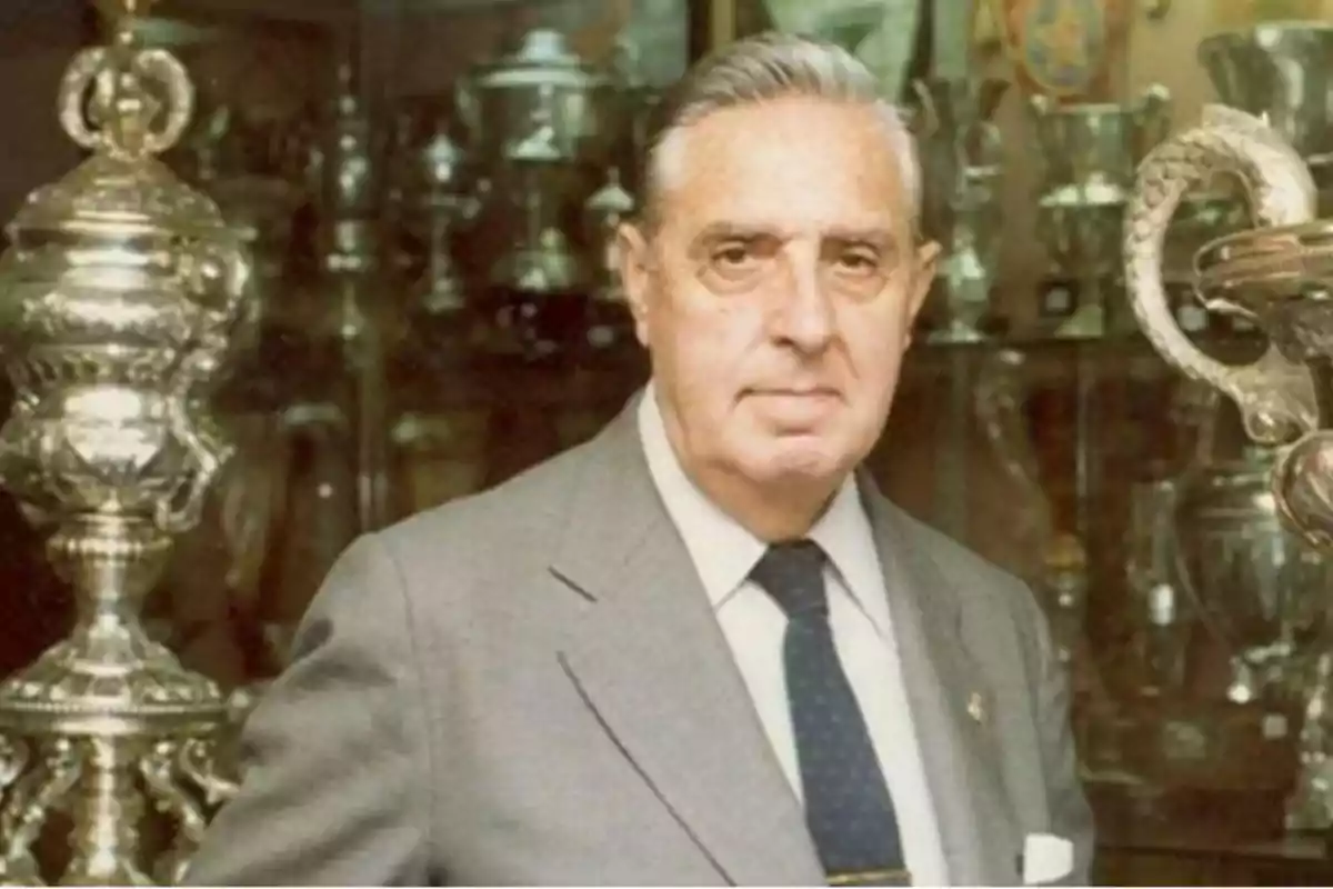 Hombre de cabello canoso con traje y corbata posando frente a una vitrina llena de trofeos plateados Hombre de cabello canoso con traje y corbata posando frente a una vitrina llena de trofeos plateados