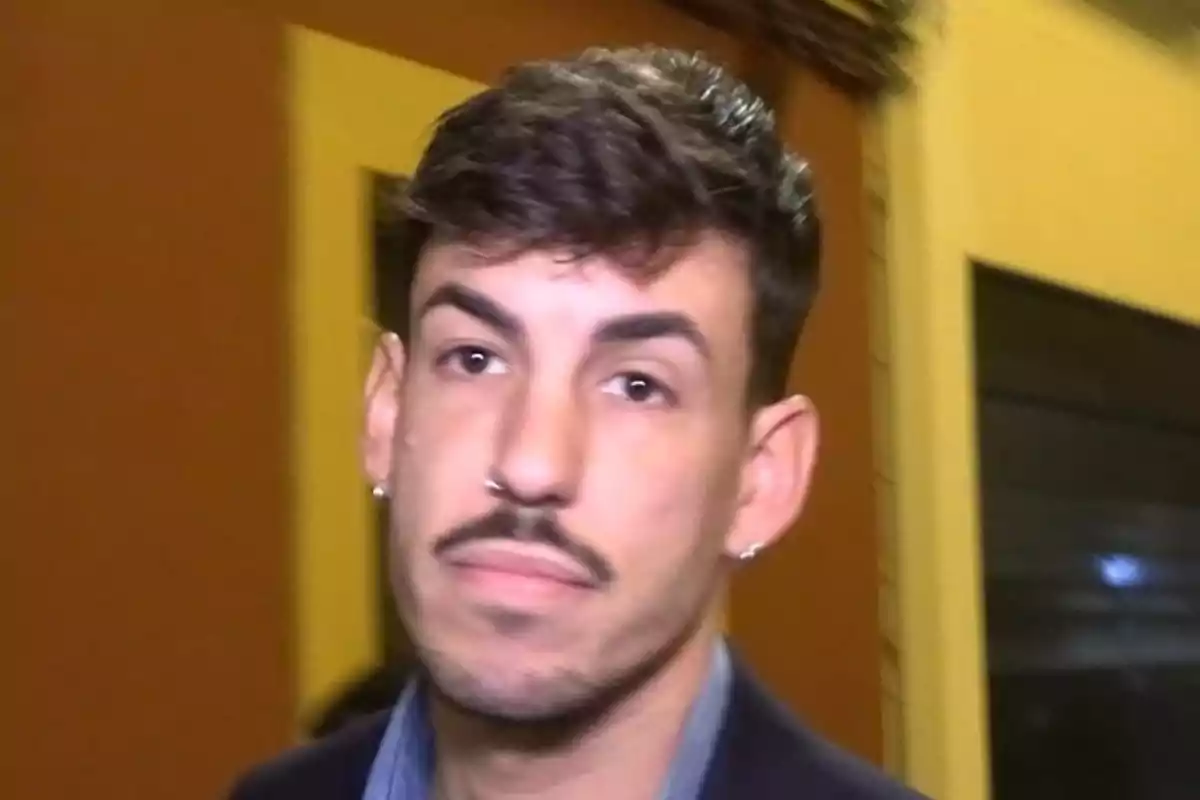 Un hombre con barba y bigote, con un aro en la nariz y aretes, mira hacia la cámara en un entorno interior con paredes de color amarillo y marrón. Un hombre con barba y bigote, con un aro en la nariz y aretes, mira hacia la cámara en un entorno interior con paredes de color amarillo y marrón.