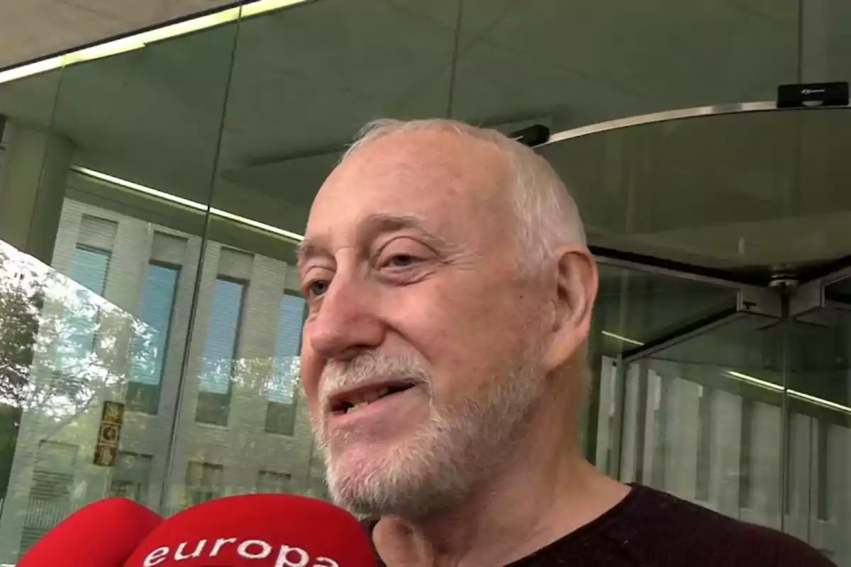 Un hombre mayor con barba blanca hablando frente a un micrófono rojo de "europa" en un entorno urbano con edificios y vidrio.