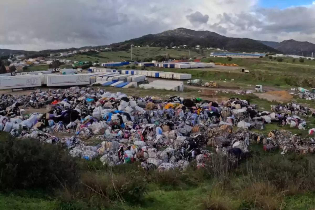 Montones de ropa desechada en un vertedero al aire libre con contenedores y montañas al fondo.