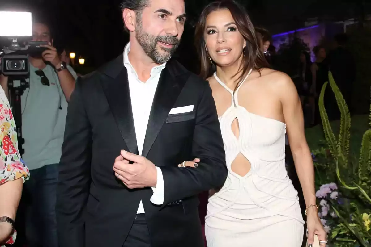 Pareja elegantemente vestida caminando en un evento nocturno rodeados de otras personas y cámaras Pareja elegantemente vestida caminando en un evento nocturno rodeados de otras personas y cámaras