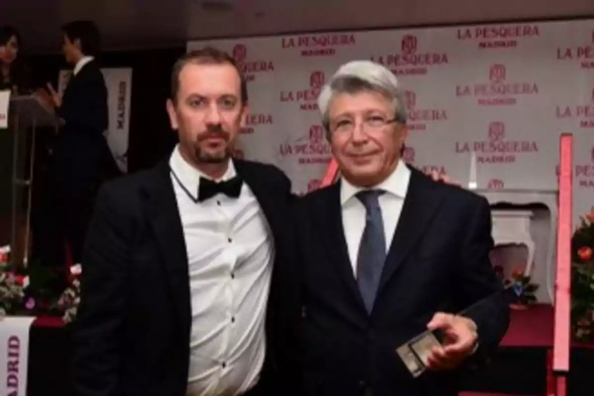 Dos hombres posan juntos en un evento formal en La Pesquera Madrid, uno de ellos sostiene un trofeo.