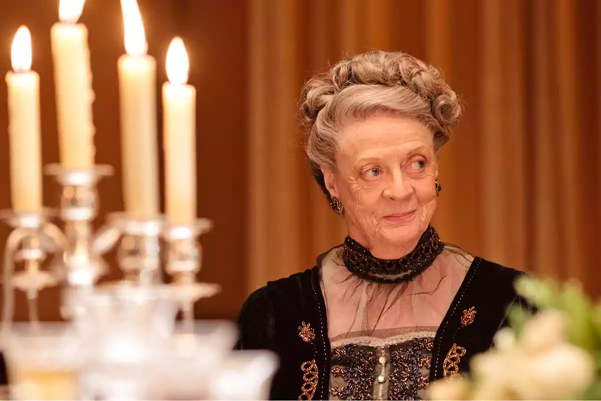 Una mujer mayor elegantemente vestida con un peinado recogido y un vestido negro adornado, sentada junto a un candelabro con velas encendidas.