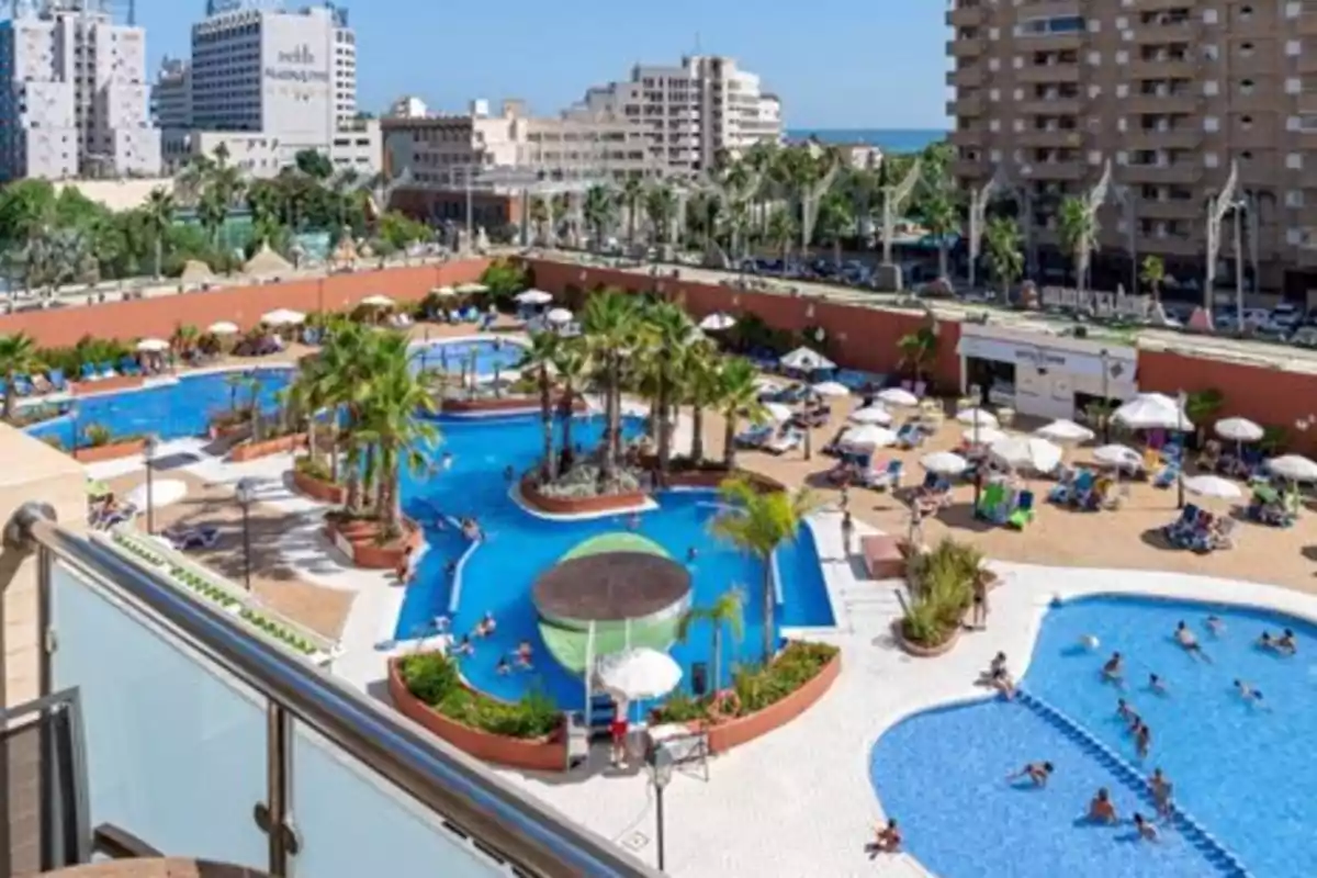 Vista aérea de un complejo hotelero con varias piscinas rodeadas de palmeras y tumbonas, ubicado en una zona urbana con edificios altos al fondo.